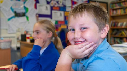 Boy and a girl in a classroom setting, the boy in the foreground smiles for the camera